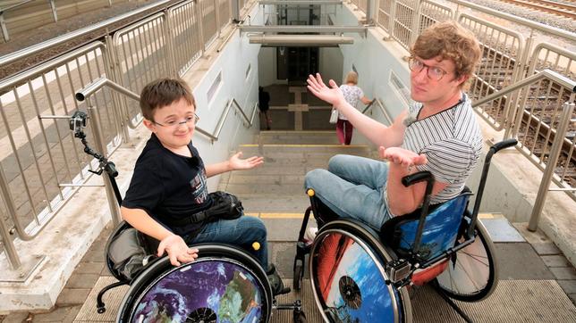Robert und Timon (l) stehen vor den Treppenstufen am Troisdorfer Bahnhof. Beide sitzen im Rollstuhl – Timon seit er 3 Jahre alt ist, Robert nur heute.
