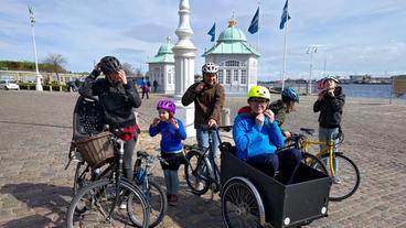 Ralph Caspers wird von seiner Gastfamilie in Kopenhagen per Fahrrad abgeholt – sie machen sich auf den Weg durch Kopenhagen, Ralph darf im Kasten des Lastenfahrrads Platz nehmen.