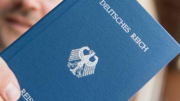 In einer Hand wird ein Heft mit dem Aufdruck «Deutsches Reich Reisepass» gehalten.