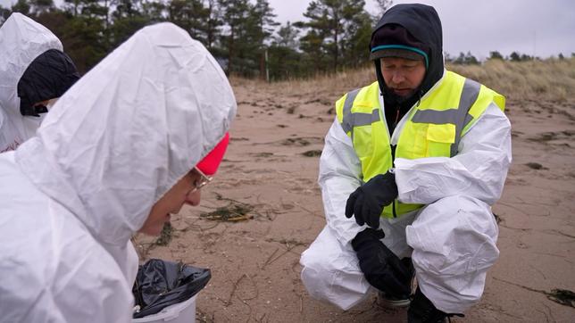 Drei Menschen in Schutzanzügen am Strand.