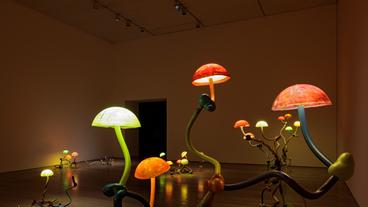 Skulpur mit mehreren pilzförmigen leuchtenden Lampenschirmen in einem abgedunkelten Raum.