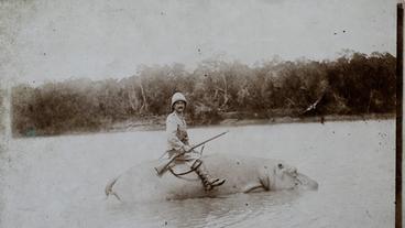 Schwarz-weiß Foto eines Mannes mit Tropenhelm und Gewehr, der auf einem im Wasser stehenden Flusspferd sitzt.