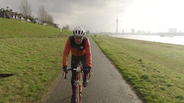 Rennradfahrer mit weißem Helm auf Radweg am Ufer eines Flusses.