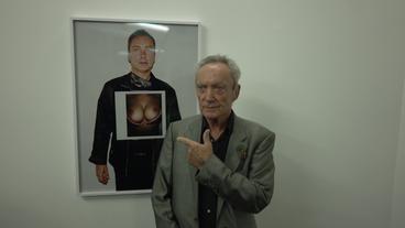 Udo Kier zeigt auf ein Foto von sich selbst mit weiblichen Brüsten.