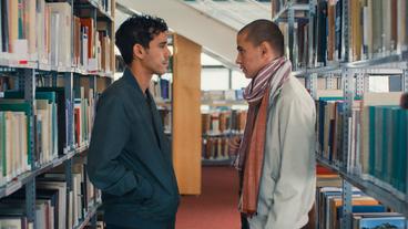 Zwei Männer stehen sich zwischen den Regalen einer Bibliothek gegenüber. 