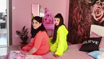 Zwei Frauen mit schwarzen langen Haaren sitzen in grellen Bademänteln auf einem Bett. Das ganze Zimmer ist rosa.