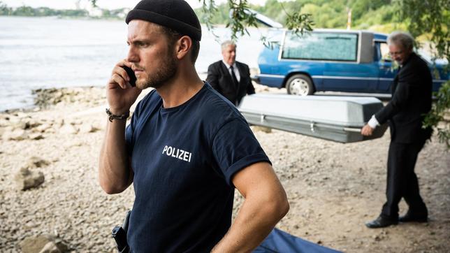 Frank van Dijk (Niklas Osterloh) telefoniert, währen die Leiche abtransportiert wird (mit Komparsen).