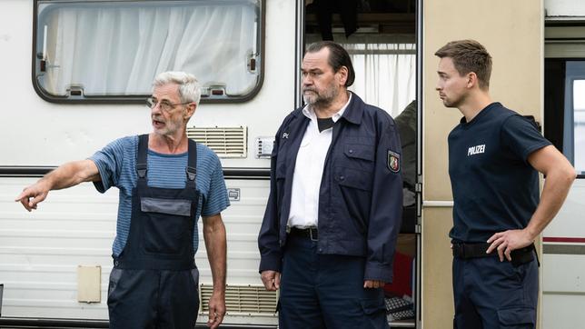 Platzwart Heinz Grünwald (Rolf Berg) berichtet Jäger (Markus John) und Mark (Timmi Trinks) von den Vorkommnissen auf dem Campingplatz.